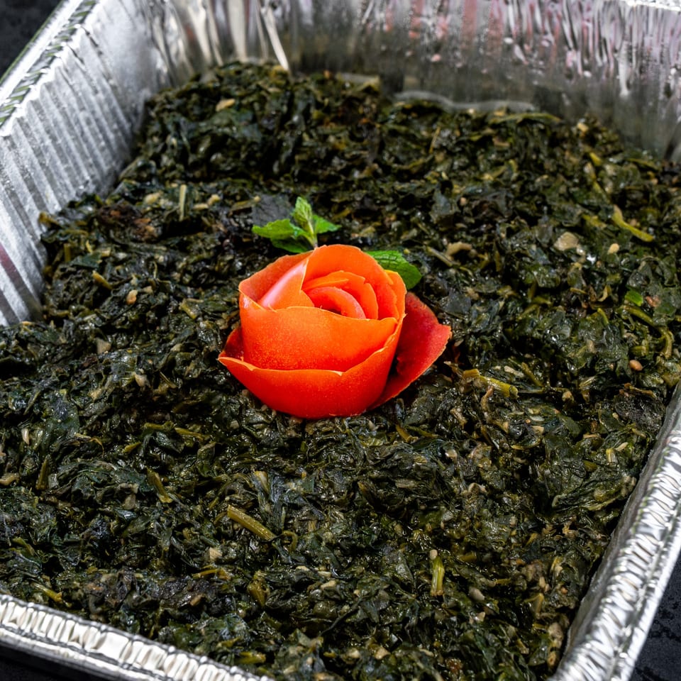 A foil tray filled with cooked green spinach, garnished with a tomato carved into a rose and mint.