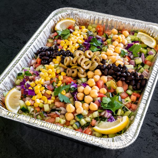 Colorful chopped salad with corn, black beans, chickpeas, olives, and lemon wedges in a foil pan.