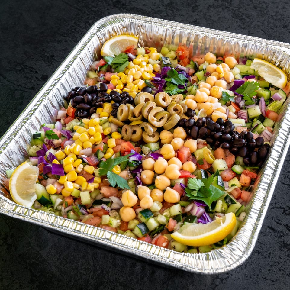 Colorful chopped salad with corn, black beans, chickpeas, olives, and lemon wedges in a foil pan.
