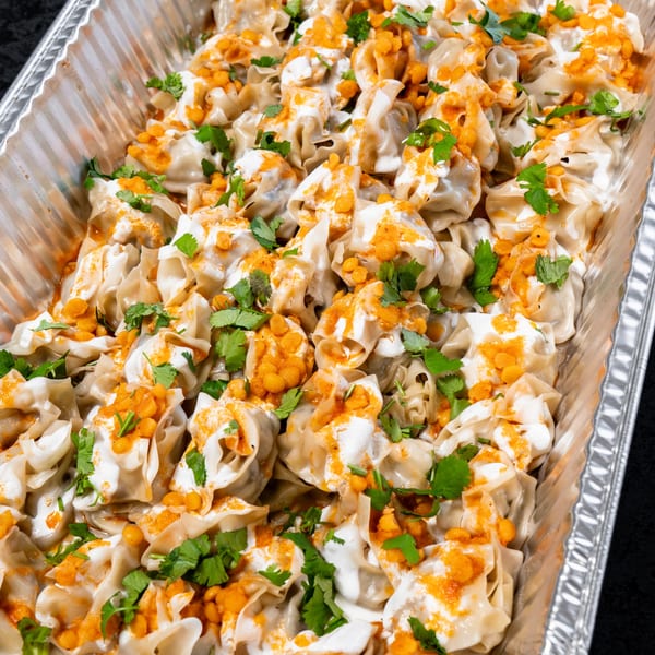 A catering tray filled with dumplings topped with white sauce, orange lentils, and fresh cilantro.