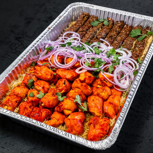 A catering tray filled with chicken tikka, seekh kebabs, rice, red onions, and cilantro.