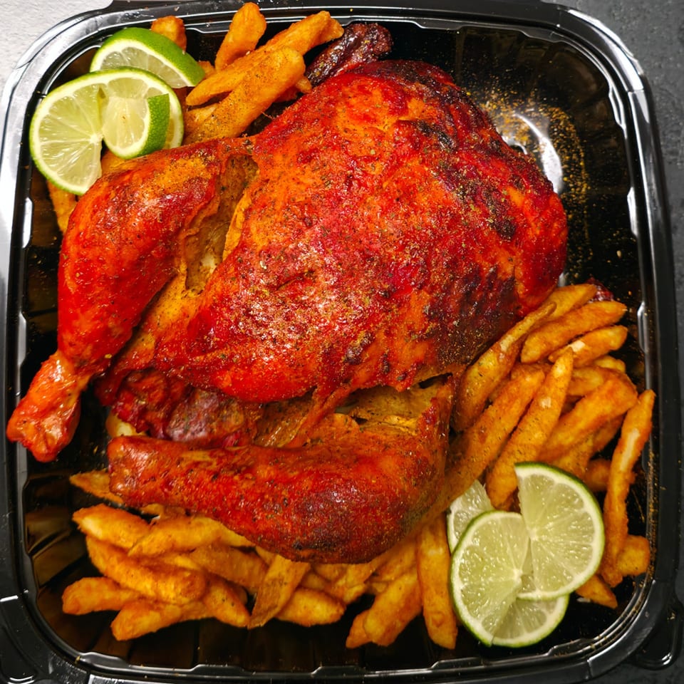 Spicy whole roasted chicken with crispy fries and lime wedges in a black takeout container.