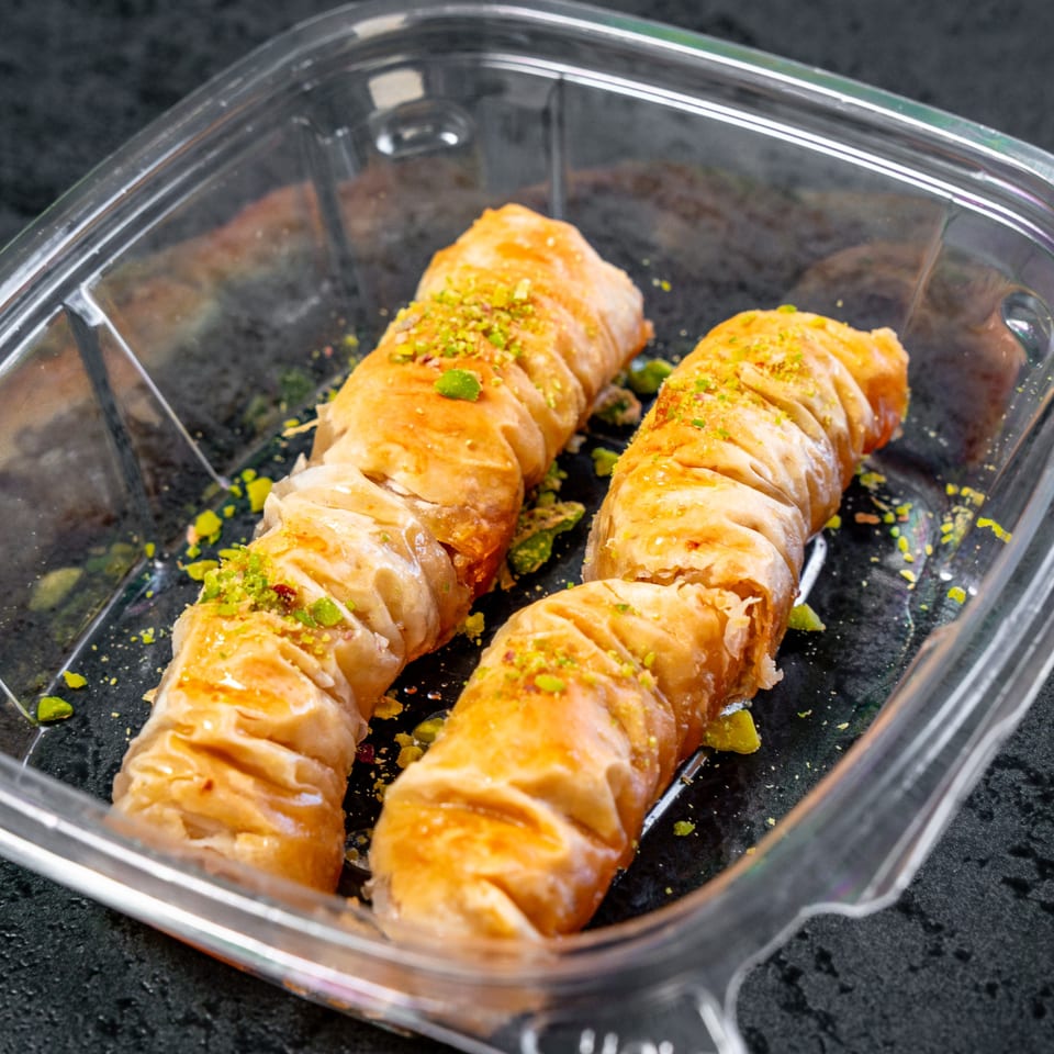 Four golden-brown baklava rolls, sprinkled with green pistachios, in a clear plastic container.